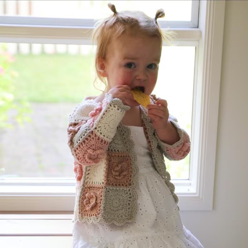 Young child wearing an open pastel granny square cardigan over a white dress.