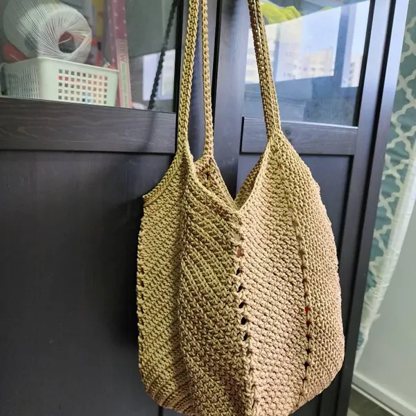 Beige crochet tote bag with long handles hanging against a dark cabinet.