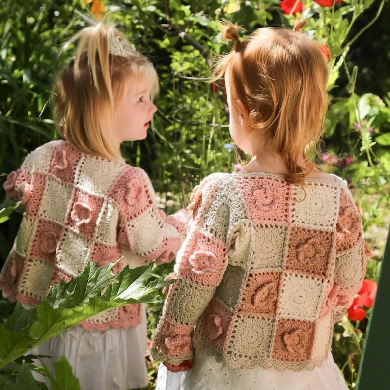 Two children wearing pastel flower granny square cardigans in a garden.