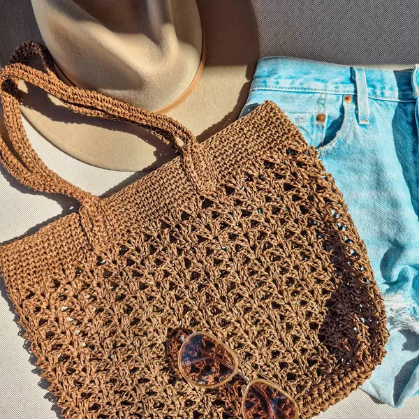 Brown openwork raffia crochet beach bag beside a hat and denim shorts.