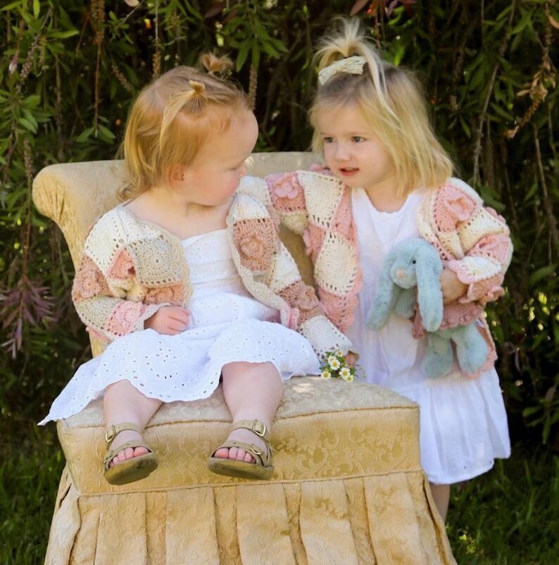 Two children seated together wearing cream and pastel floral crochet cardigans.